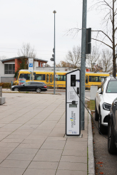 Erkennbar ist eine Ladestation für Elektromobilität. Rechts daneben sieht man zwei Autos. Im Hintergrund ist die UBahn abgebildet, die vorbeifährt.
Elektromobilität